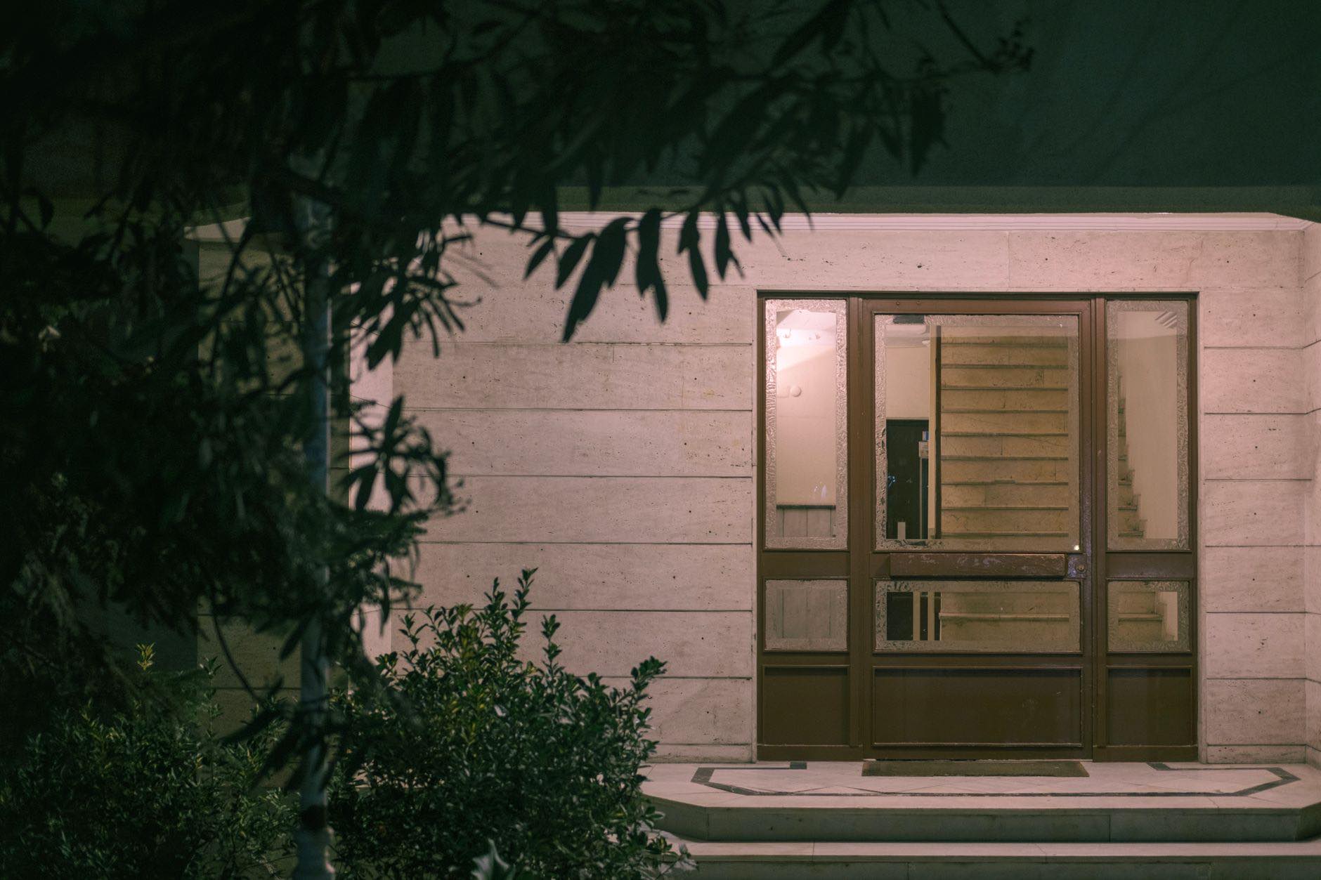 A lit doorway at the base of a dark building at night, seen past overhanging branches